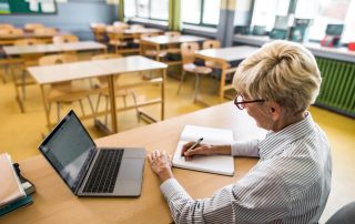 A teacher sitting in an empty classroom with desks. Working at desk taking notes at her laptop.