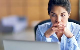 A woman sitting at a desk looking at a laptop with her hands clasped