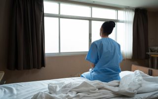 The back of a woman sitting on the edge of bed at a psychiatric facility looking out a window.