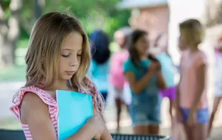 A young girl standing alone at school with blurred image of girls talking in the background