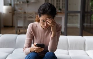 A woman sitting on a couch in a dark home staring concerned looking at her phone with hand on her head