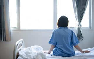 The back of a woman patient sitting on the edge of a hospital bed looking out a window