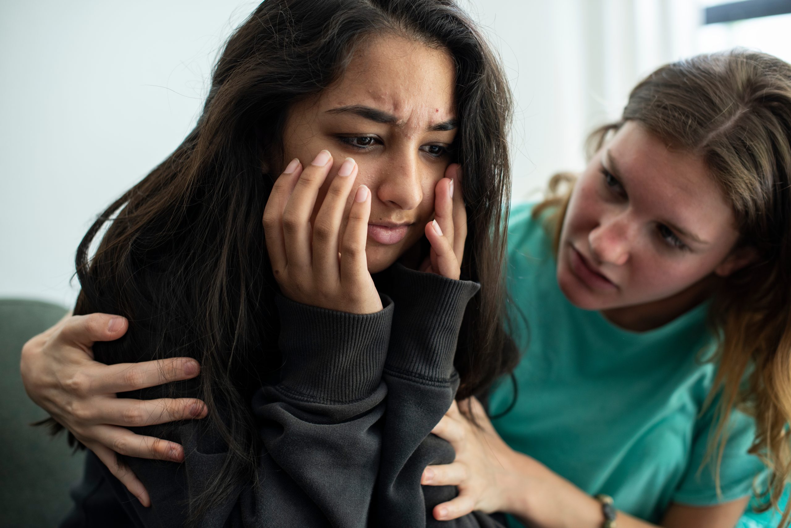 Two young females sitting together. One is distressed with her face in hands and her friend is comforting her.