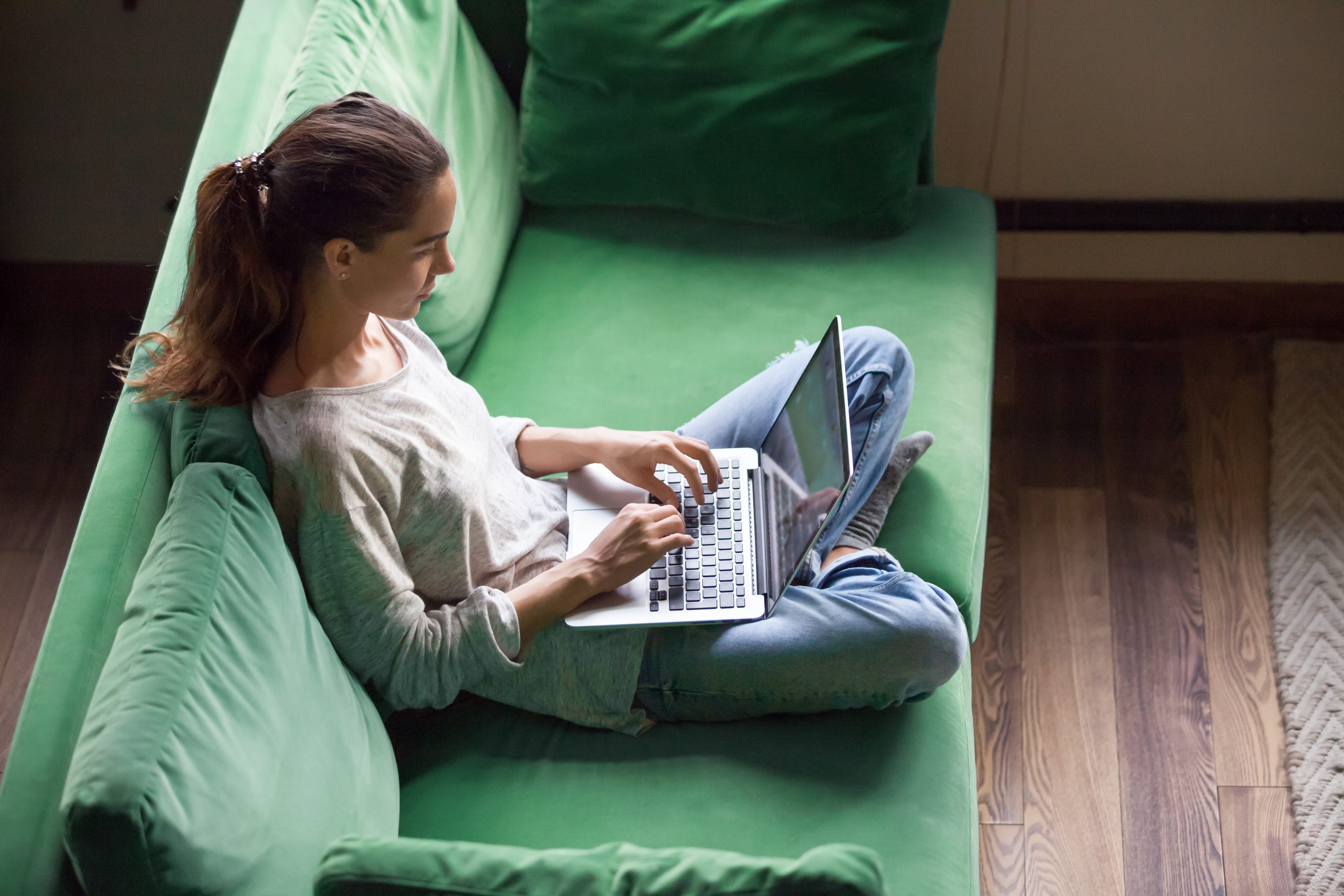 A young woman sitting alone on a green couch at home using a laptop.