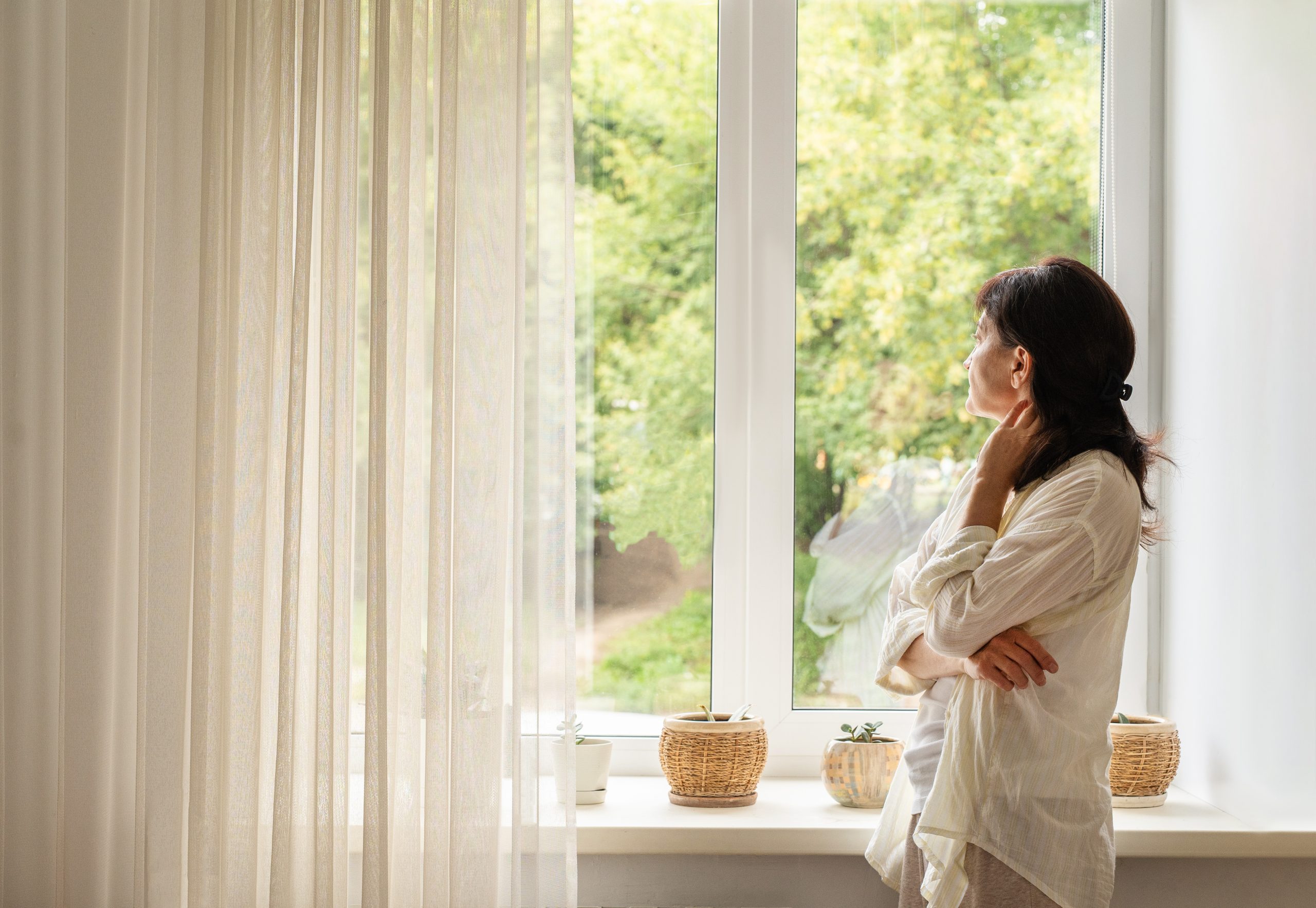 Mature woman in a white blouse stands by large, light-filled window with sheer curtains, gazing at green trees. She feels at peace, experiencing quiet solitude.